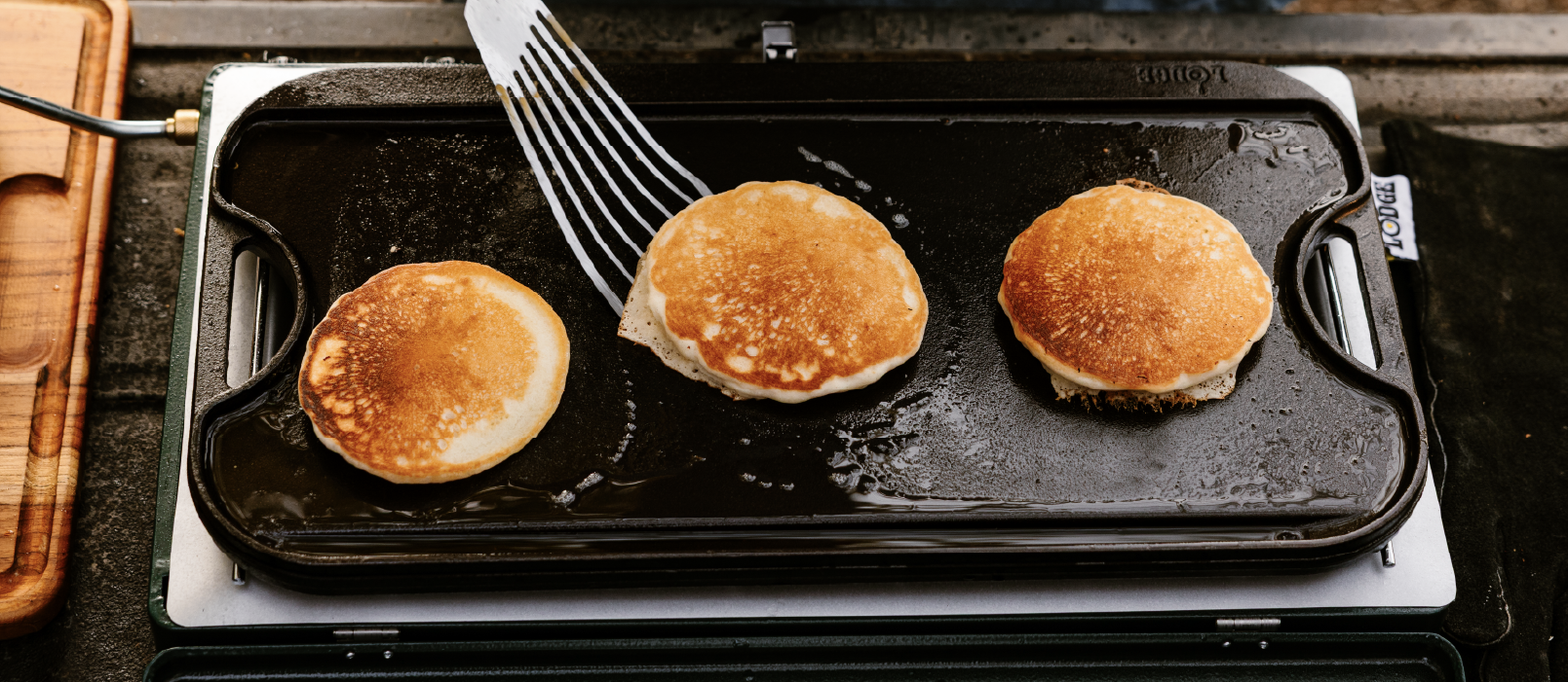 Three golden-brown pancakes cooking on a black flat-top griddle with a metal spatula being used to flip pancake, set on a white portable burner outside.