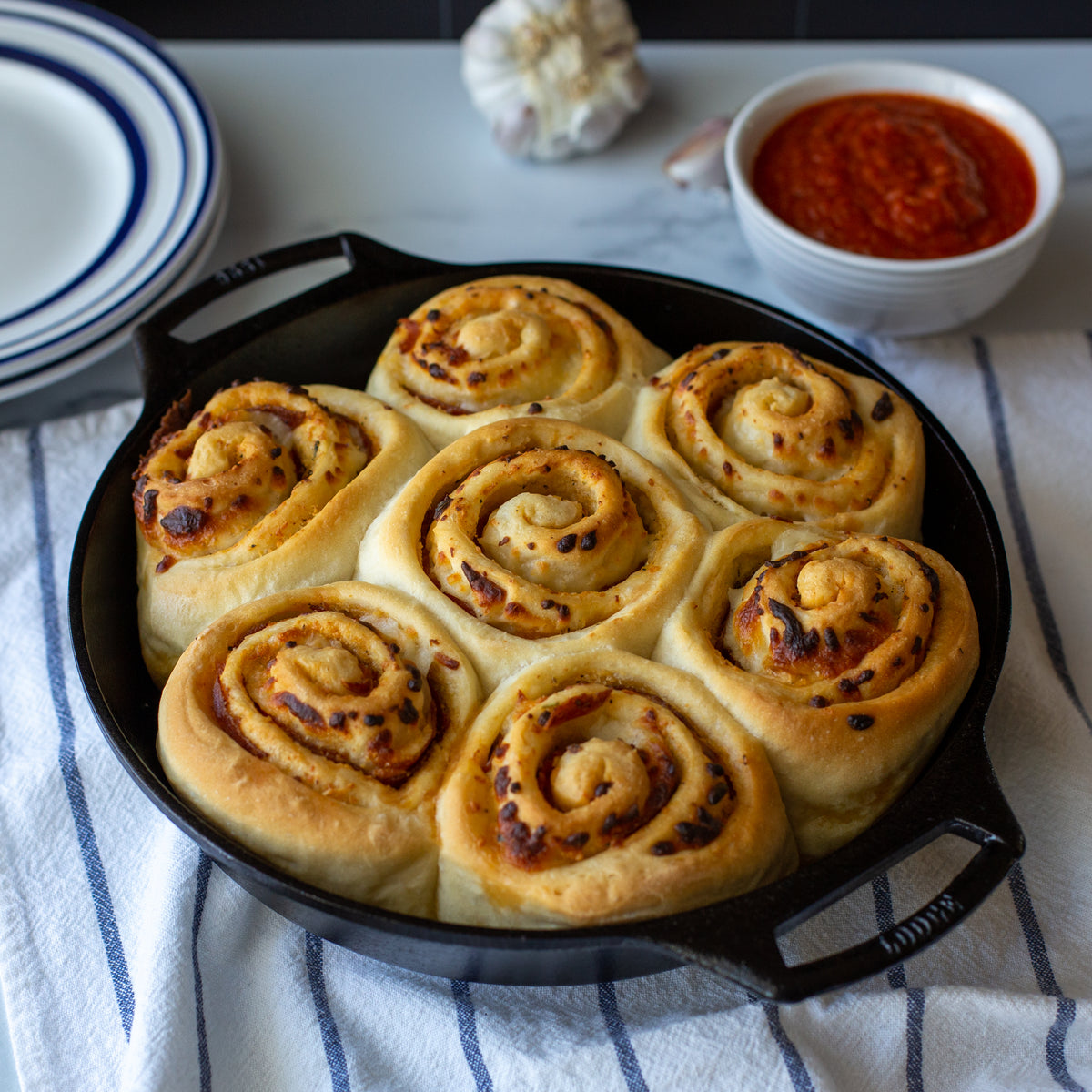Seven baked pizza rolls in a black cast iron skillet, set on a white and blue striped kitchen towel with a bowl of marinara sauce and garlic in the background.