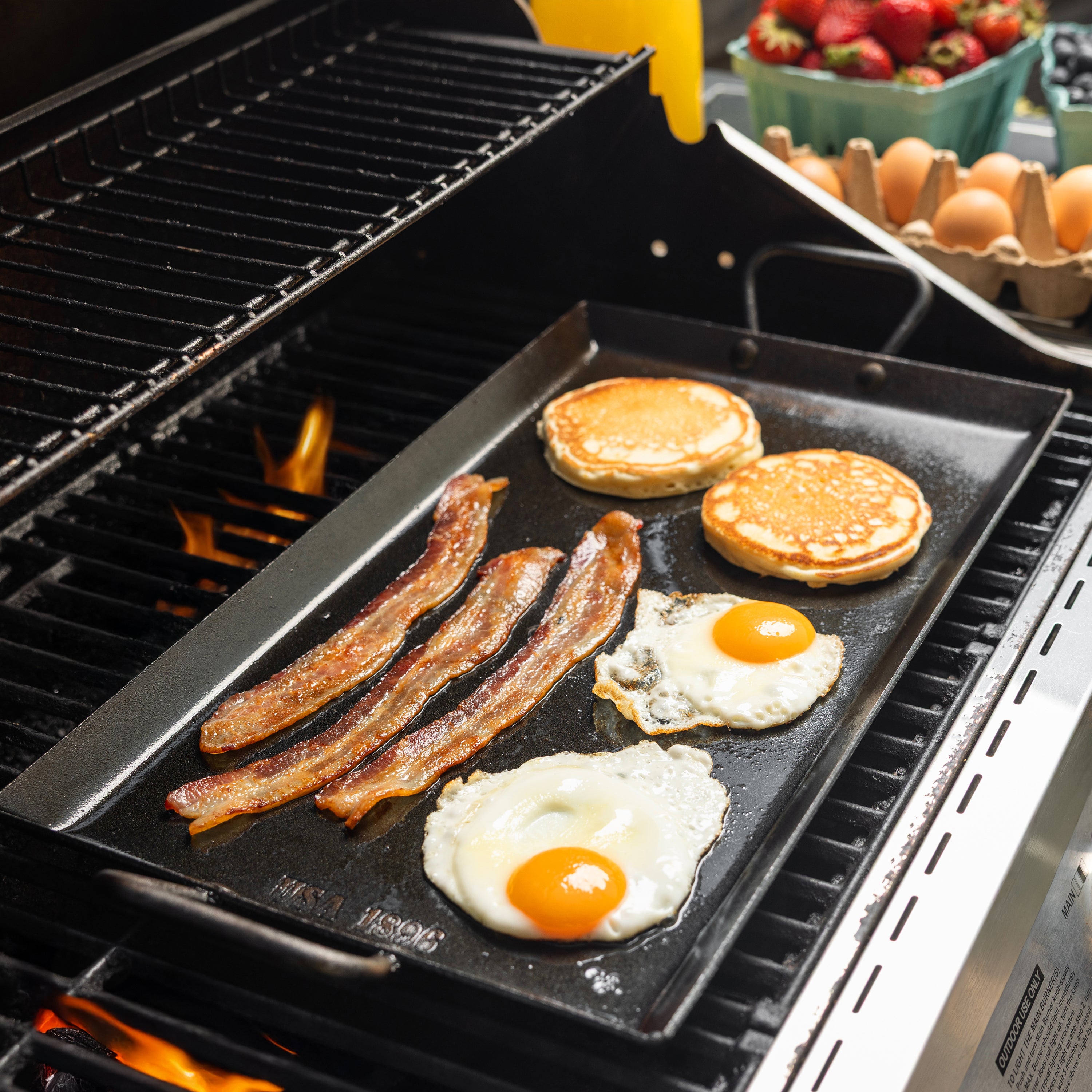 A black flat-top carbon steel griddle cooking pancakes, bacon, eggs, and a sunny-side-up egg on a grill, with a carton of eggs and strawberries visible in the background.
