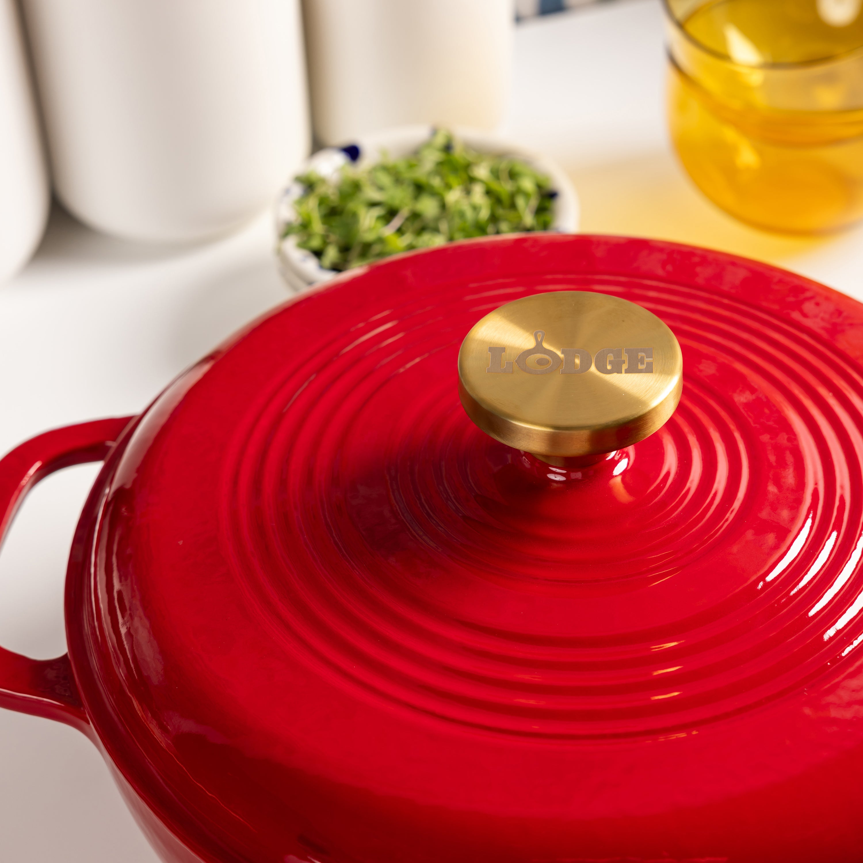 A close-up of a red Lodge enameled cast iron Dutch oven with a gold-toned lid knob, set on a white countertop with blurred kitchen items in the background.