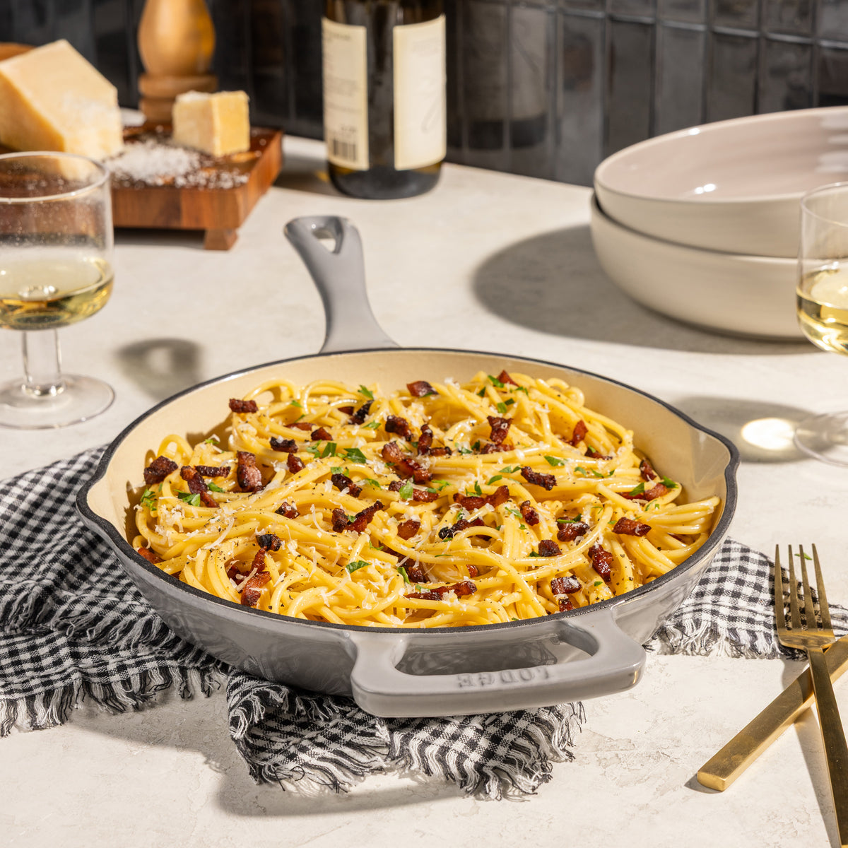 A cast iron skillet with a cream interior, shown filled with carbonara pasta and garnished with crispy bacon bits and parmesan cheese. The skillet is set on a black and white checkered cloth on a light stone countertop, with dining props like a wine bottle, glasses, and plates in the background.