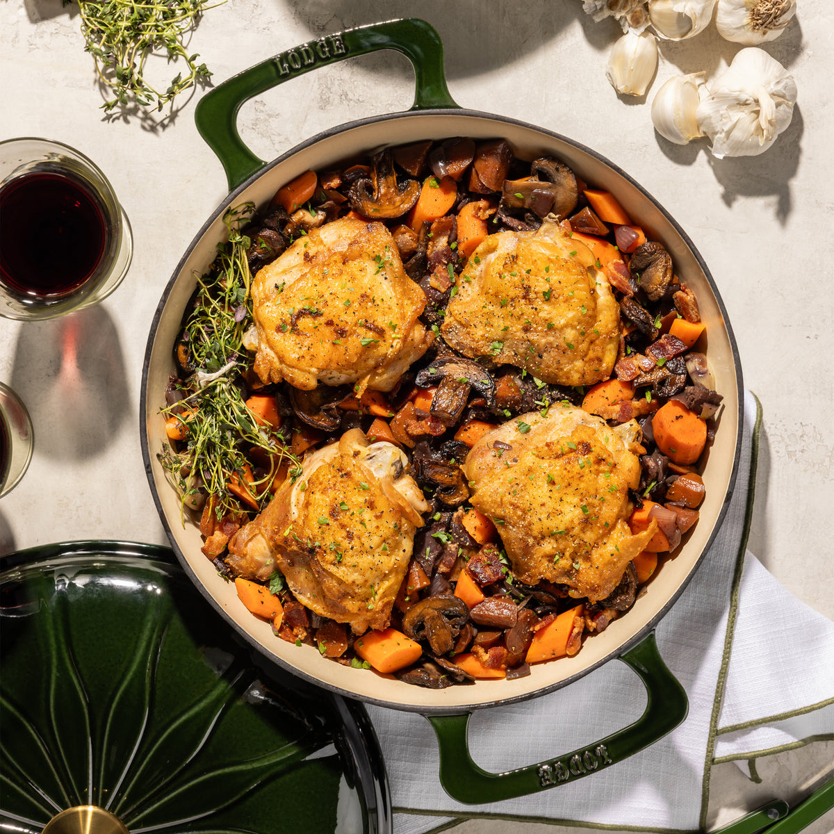 A top-down view of a green enameled cast iron Dutch oven filled with roasted chicken, carrots, and mushrooms, styled on a light stone surface with garlic, a glass of red wine, and herbs.