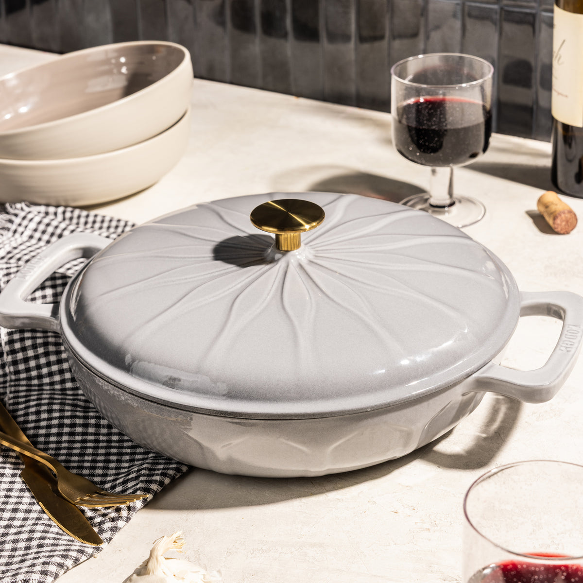 A light gray enameled cast iron casserole dish with a matching lid, featuring a gold-toned knob, styled on a kitchen counter with wine glasses, bowls, and gold cutlery.
