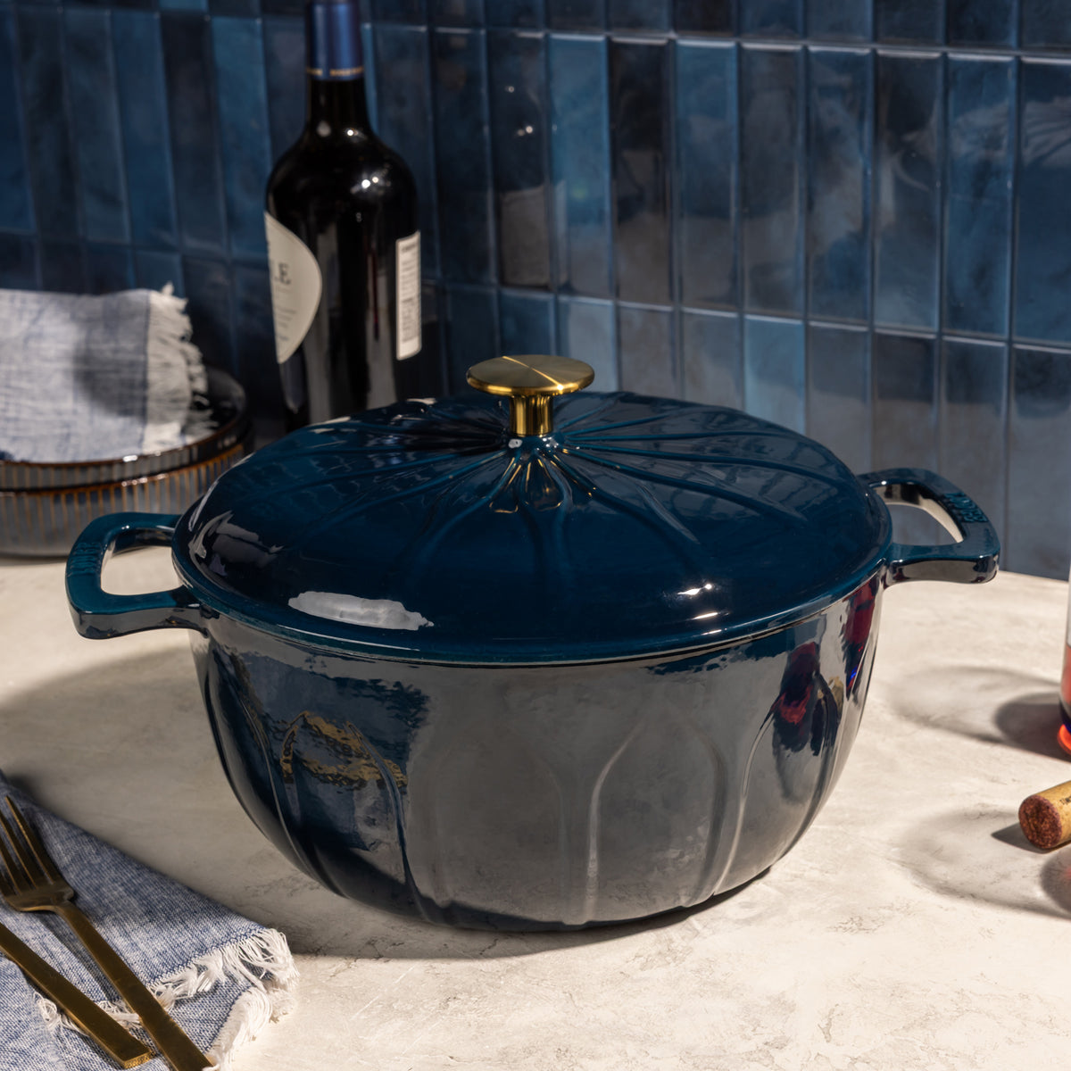 A dark blue enameled cast iron Dutch oven with a lid and gold-tone knob, set on a light stone countertop with a blue tiled backsplash, a wine bottle, and dining props in the background.