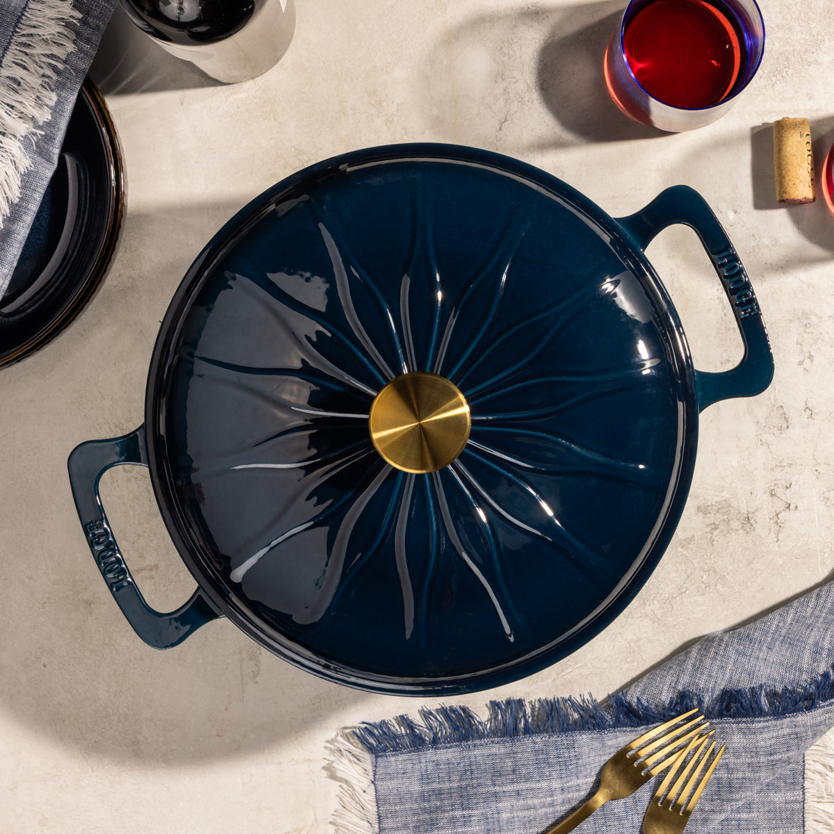 A top-down view of a dark navy blue enameled cast iron pot with a matching lid featuring a sunburst design and a gold center knob, styled on a light stone surface with dining props.