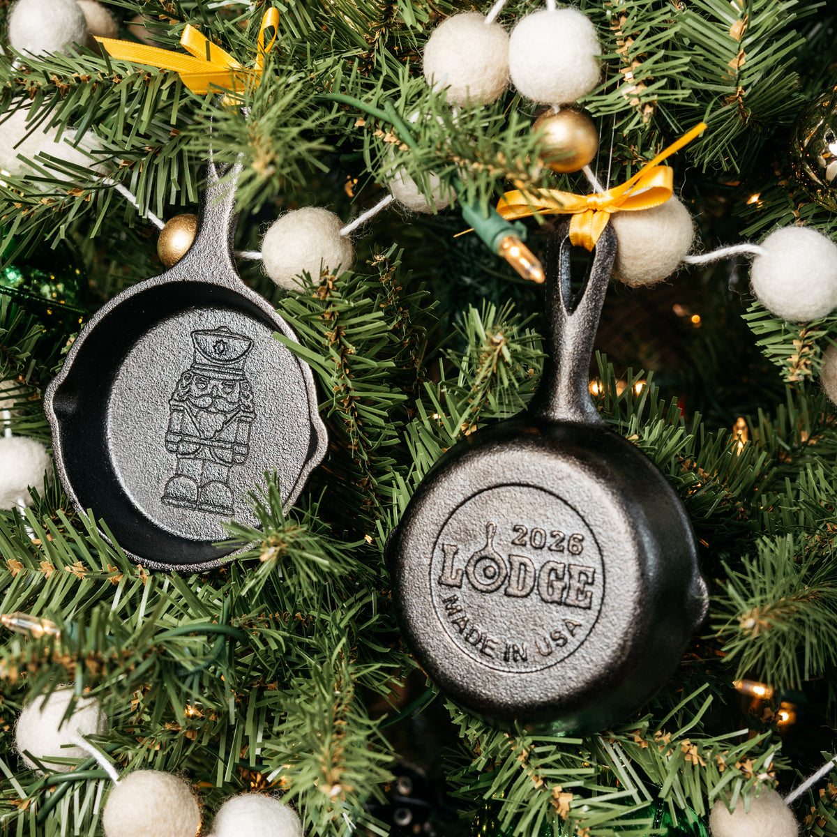 Two cast iron skillet-shaped Christmas ornaments hanging on a decorated Christmas tree. One ornament is shown from the back with the embossed Lodge logo and '2026 Made in USA' text, and the other is shown from the front with an embossed nutcracker design.