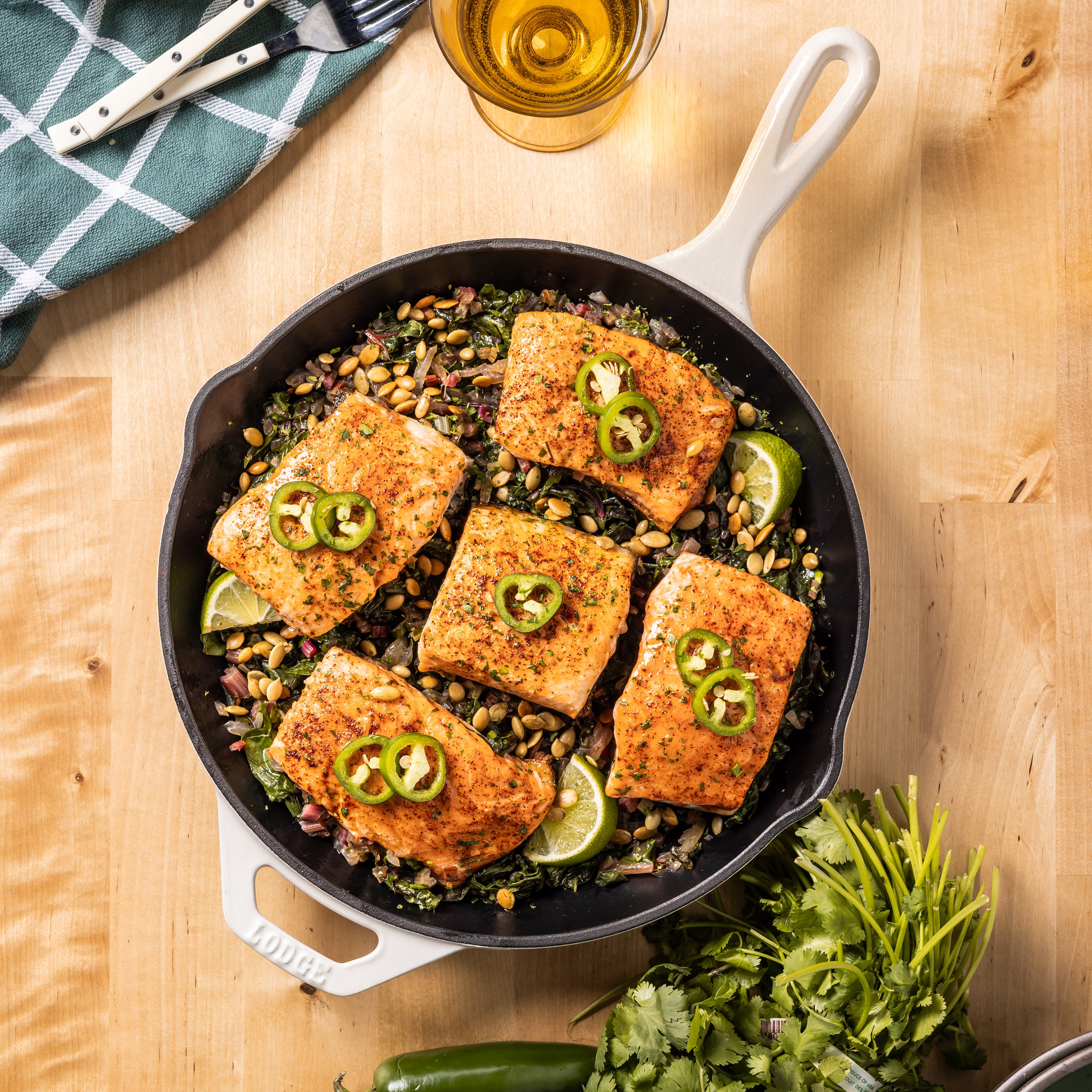 A cast iron skillet with white handles sits on a wooden table, filled with seasoned fish fillets topped with sliced jalapeño and lime, served over a bed of greens and nuts. A glass of beer, a folded napkin, and fresh cilantro are arranged around the skillet.