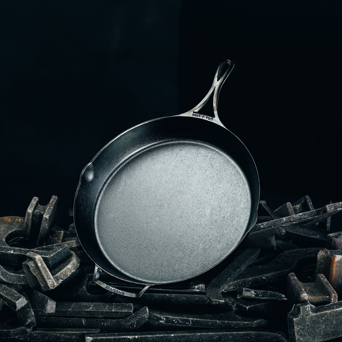 A dark cast iron skillet with a textured non-stick surface, resting on a pile of dark metal tools against a solid black background.