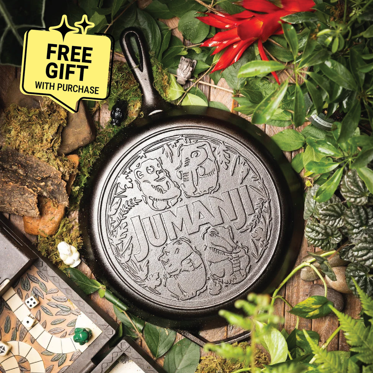 A cast iron skillet with an embossed 'JUMANA' design, surrounded by lush green foliage, moss, rocks, and a small game board on a rustic wooden surface. Featuring a badge saying "Free gift with purchase".