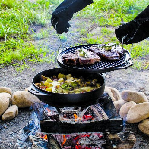 Someone using the hook and carry handles to lift grill pan/griddle after searing steak. Below the grill pan is the wok portion cooking a stew over the fire using our Fire and Cook Stand.