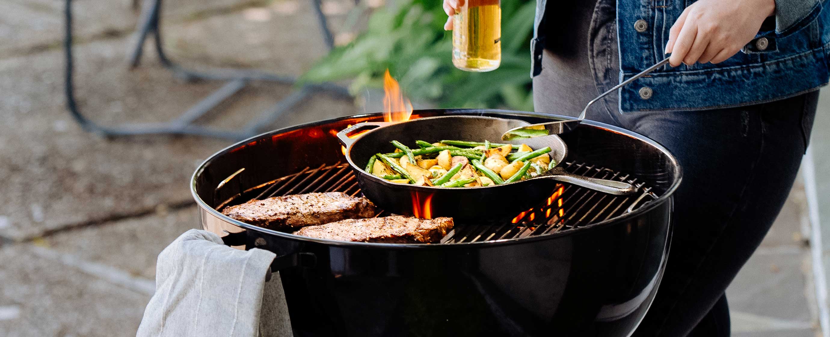 Person grilling vegetables in a cast iron skillet and meats on a barbecue.