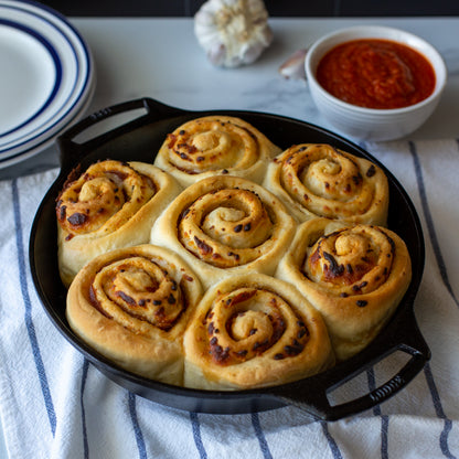 Seven baked pizza rolls in a black cast iron skillet, set on a white and blue striped kitchen towel with a bowl of marinara sauce and garlic in the background.