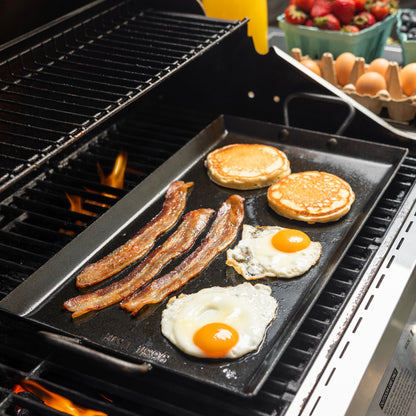 A black flat-top carbon steel griddle cooking pancakes, bacon, eggs, and a sunny-side-up egg on a grill, with a carton of eggs and strawberries visible in the background.