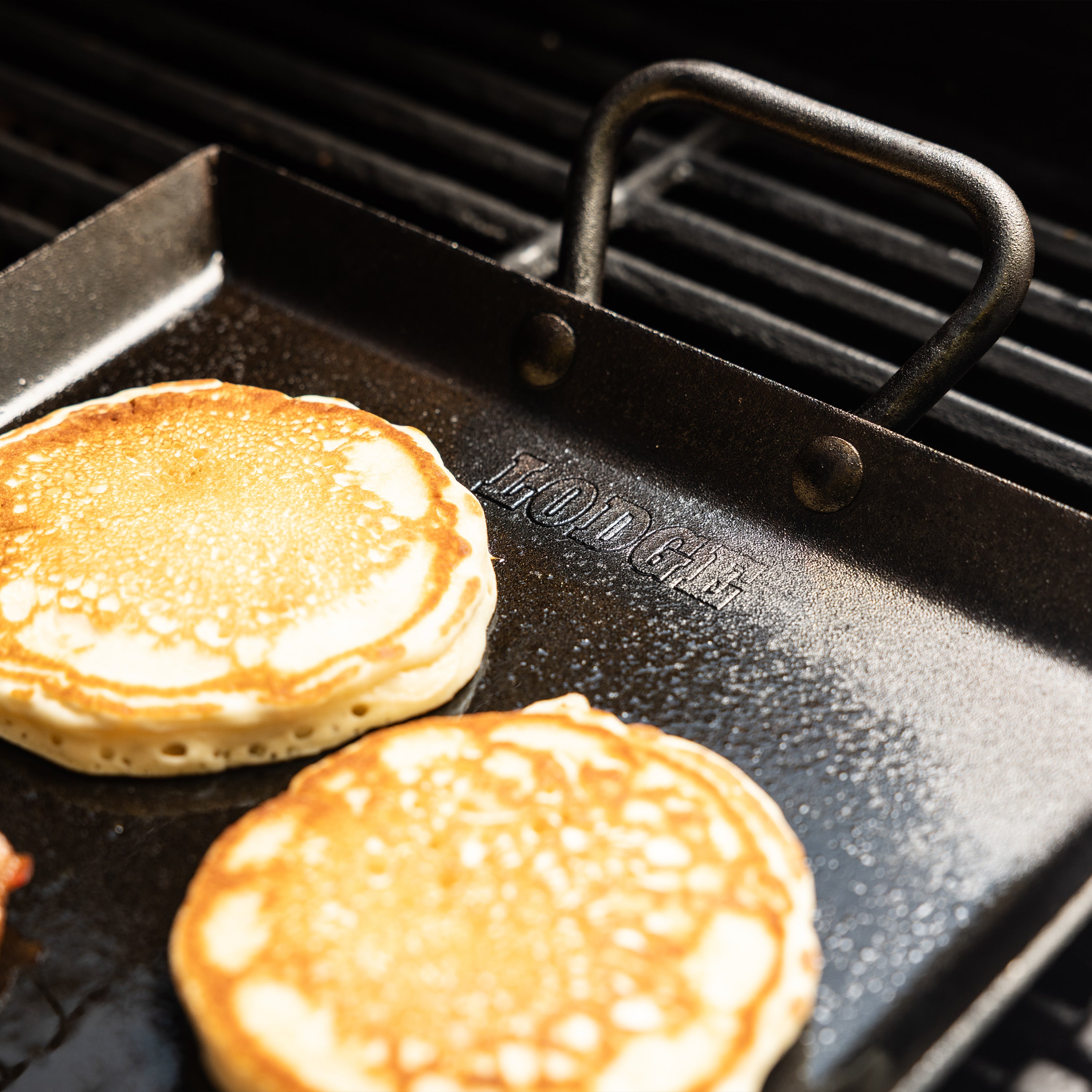 Two golden-brown pancakes cooking on a black griddle with a handle, set on a grill grate.