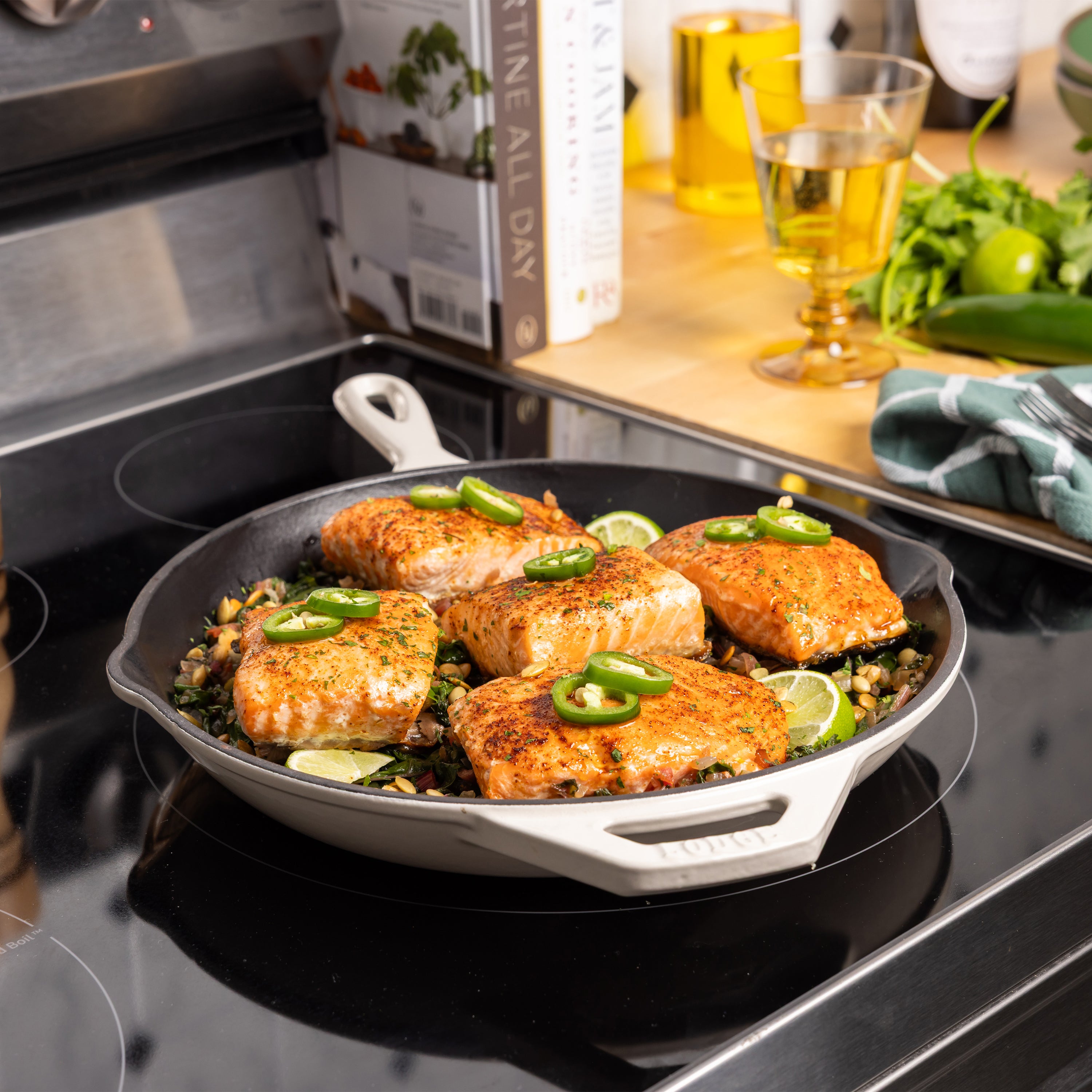 A cast iron skillet on a black induction stovetop, cooking salmon fillets topped with sliced jalapeño and lime over a green herb base. A cookbook, glass of oil, and fresh herbs are visible in the kitchen background.