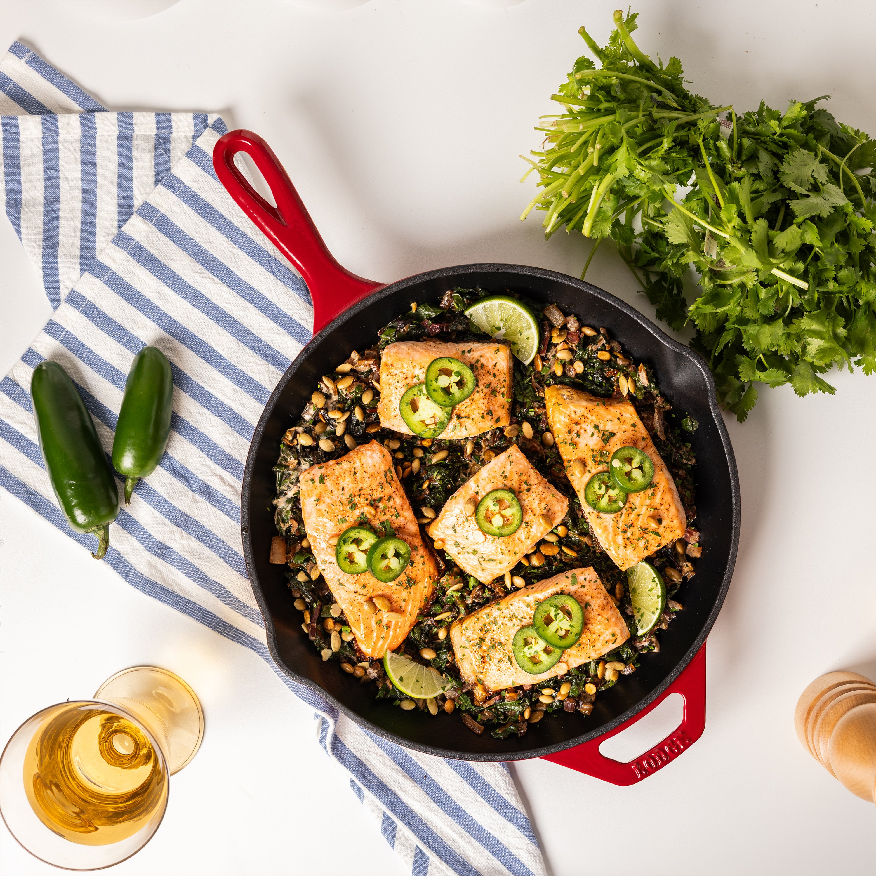 Salmon fillets with jalapeños and lime in a red skillet on a white surface with cilantro and a glass of wine.