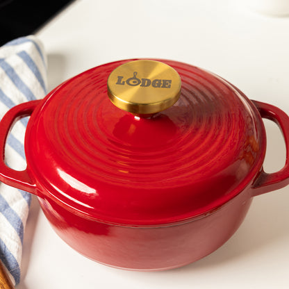 A red Lodge enameled cast iron Dutch oven with a matching lid and a gold-toned knob, shown on a white surface with a striped cloth partially visible on the left.
