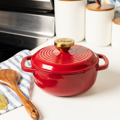 A red enameled cast iron Dutch oven with a lid and gold-tone knob, styled on a white countertop with a striped cloth, wooden spoon, and kitchen canisters in the background.