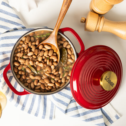 A red enameled cast iron pot filled with baked beans, shown from above on a white surface with a blue-and-white striped cloth. The wooden spoon rests inside the pot, and the matching red lid with a gold-toned knob sits beside it.