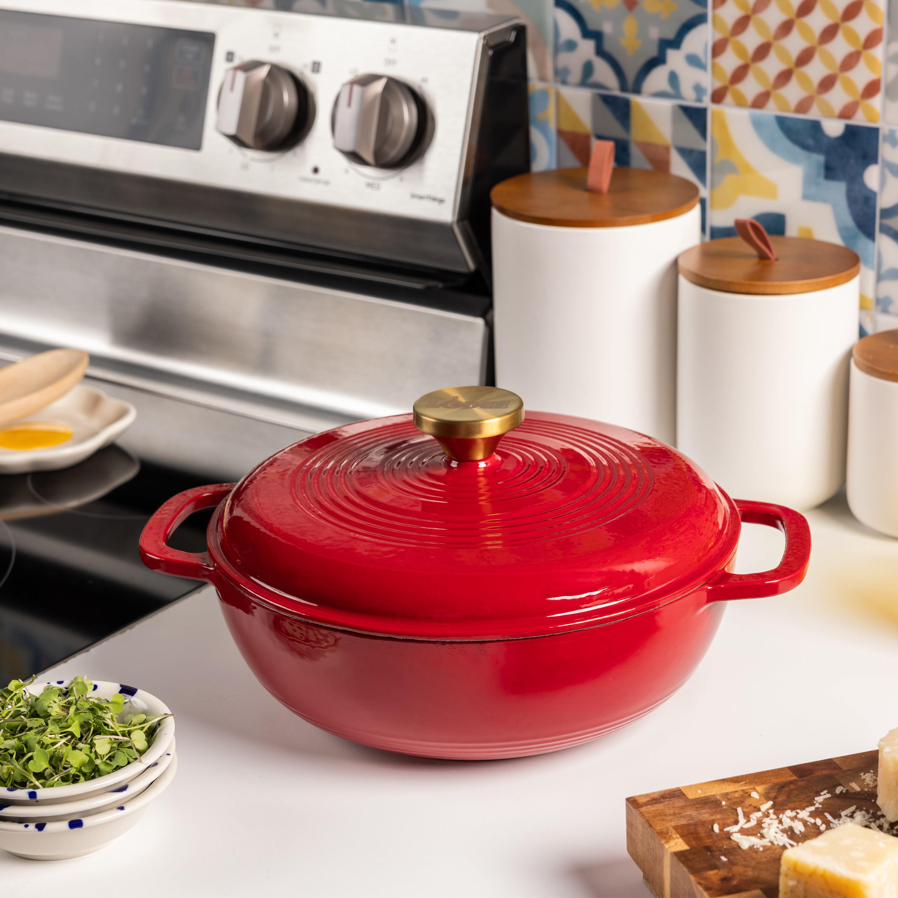 A red enameled cast iron Dutch oven with a matching lid and a brass knob, set on a white kitchen counter in front of a stainless steel stove.