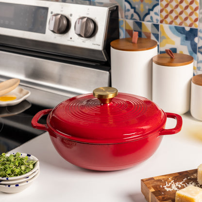 A red enameled cast iron Dutch oven with a matching lid and a brass knob, set on a white kitchen counter in front of a stainless steel stove.