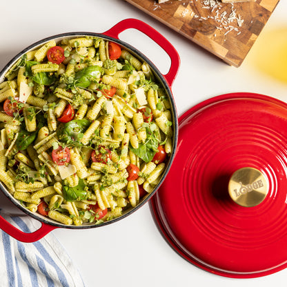 A red enameled cast iron Dutch oven with a matching lid, filled with a green pasta salad, on a white surface with a wooden cutting board in the background.