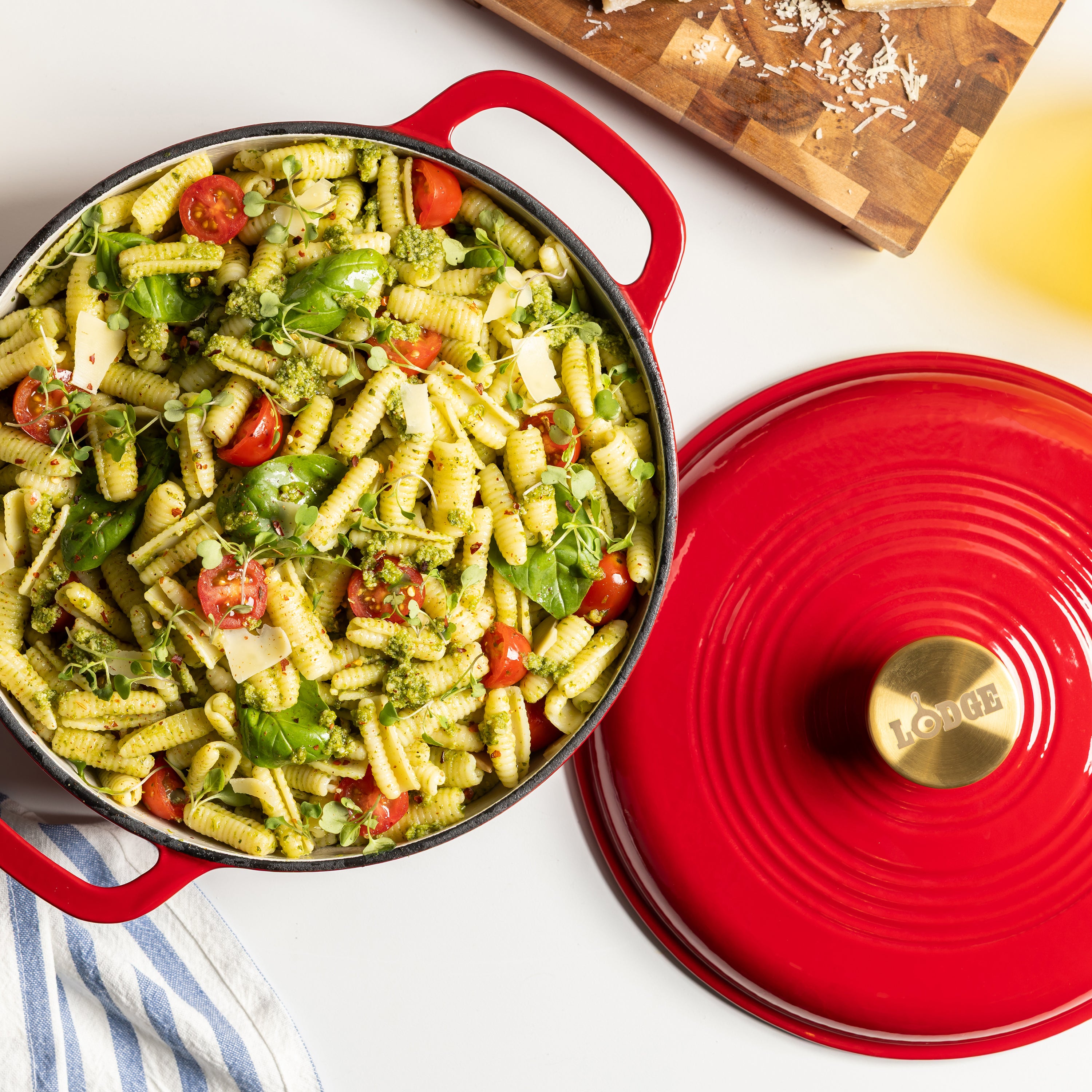 A red enameled cast iron Dutch oven with a matching lid, filled with a green pasta salad, on a white surface with a wooden cutting board in the background.