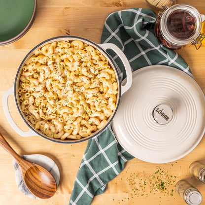 A top-down view of a creamy macaroni pasta in a white enameled cast-iron Dutch oven with a lid, placed on a light wood table with a green checkered cloth, wooden spoon, and seasoning jars.