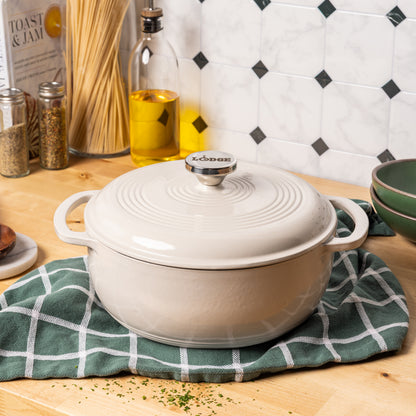 A cream-colored enameled cast iron Dutch oven with a matching lid sits on a green and white checkered kitchen towel on a wooden countertop, with kitchen props like a bottle of olive oil, pasta, and spices in the background.