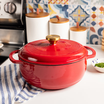 A red enameled cast iron Dutch oven with a matching lid and a brass knob, set on a white kitchen counter in front of a stainless steel stove.