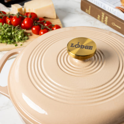 A beige Lodge enameled cast iron Dutch oven with a gold-tone knob, shown in a kitchen setting with tomatoes, cheese, and a cookbook blurred in the background.