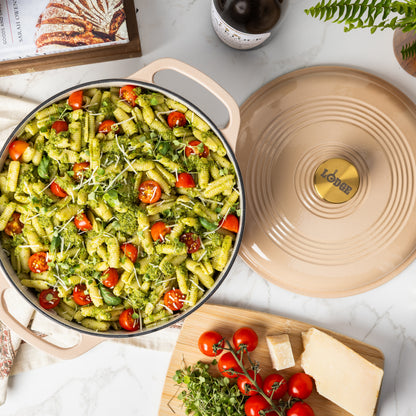A beige enameled cast iron Dutch oven filled with green pesto pasta, tomatoes, and microgreens, shown on a white marble surface with its matching lid beside it and cooking ingredients nearby.