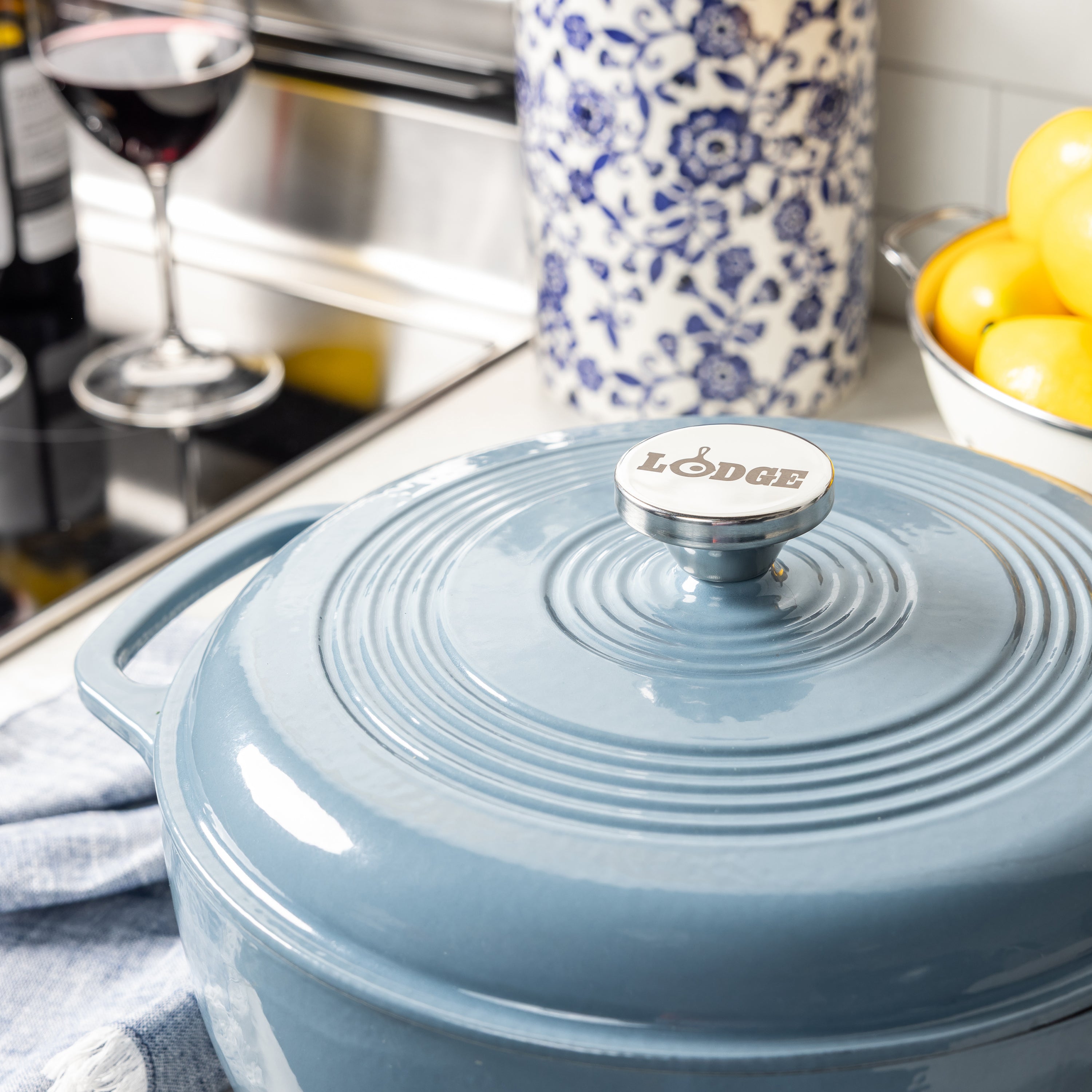 A light blue Lodge enameled cast iron Dutch oven with a matching lid, shown in a kitchen setting with a wine glass, floral jar, and lemons in the background.