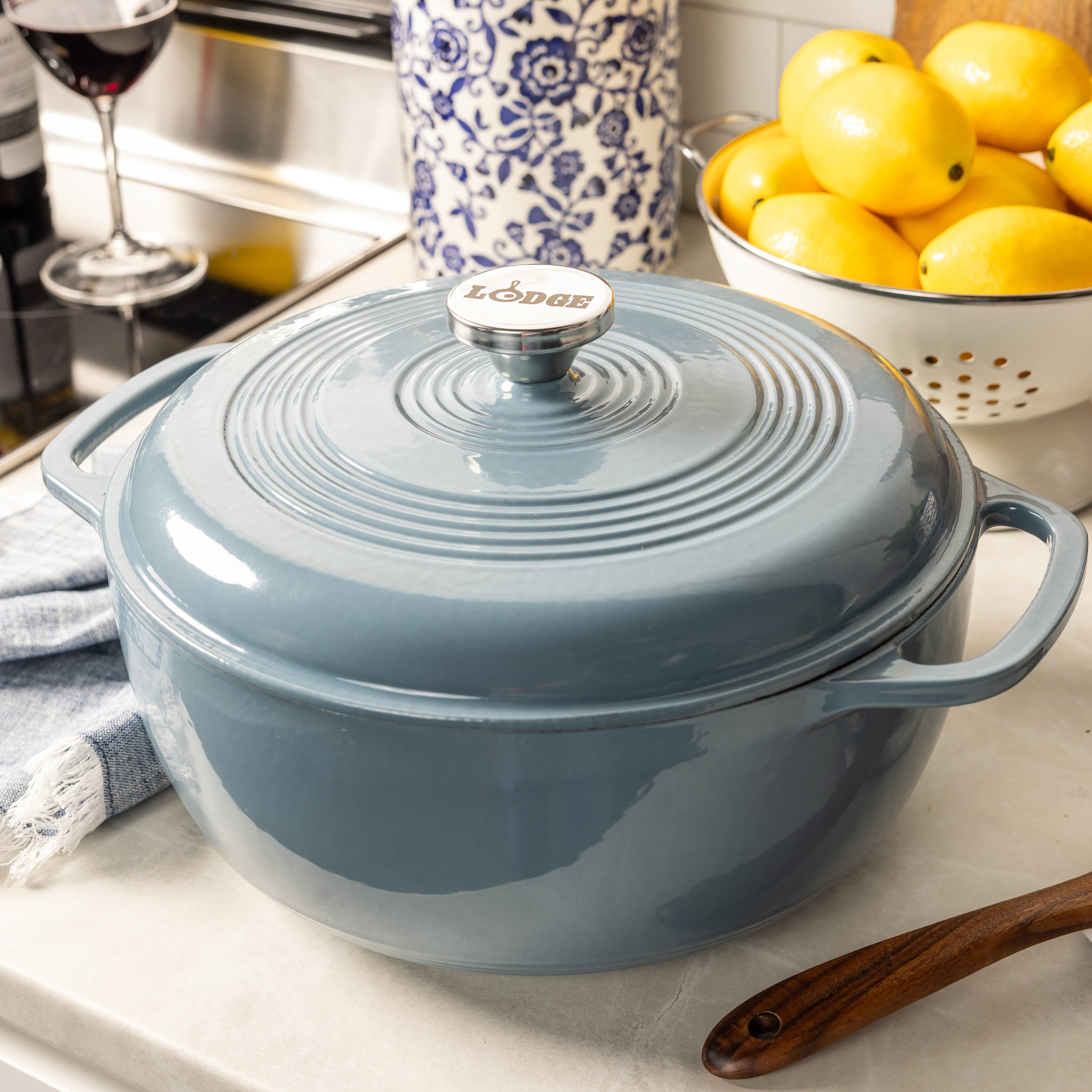 A large light blue enameled cast iron Dutch oven with a lid, shown on a kitchen counter with lemons, a wine glass, and a patterned jar in the background.