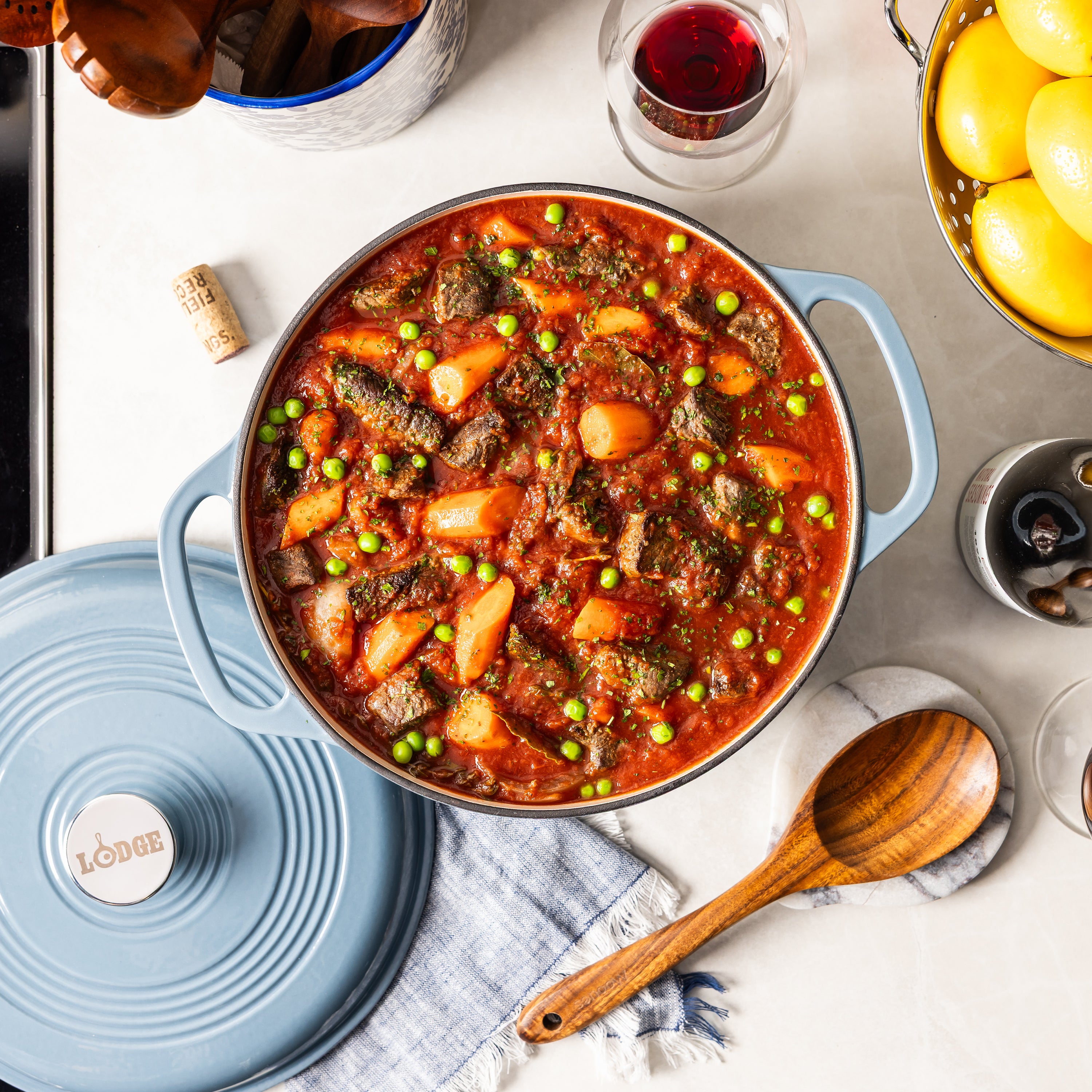 A blue Lodge enameled cast iron pot filled with a hearty beef and potato stew, shown in a styled kitchen setting with a matching lid, wooden spoon, and wine glass.