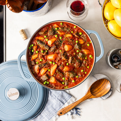 A blue Lodge enameled cast iron pot filled with a hearty beef and potato stew, shown in a styled kitchen setting with a matching lid, wooden spoon, and wine glass.
