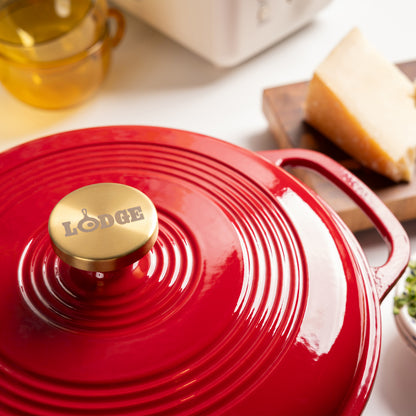 A red Lodge enameled cast iron Dutch oven with a gold-toned knob, shown close-up on a white countertop with kitchen props in the background.