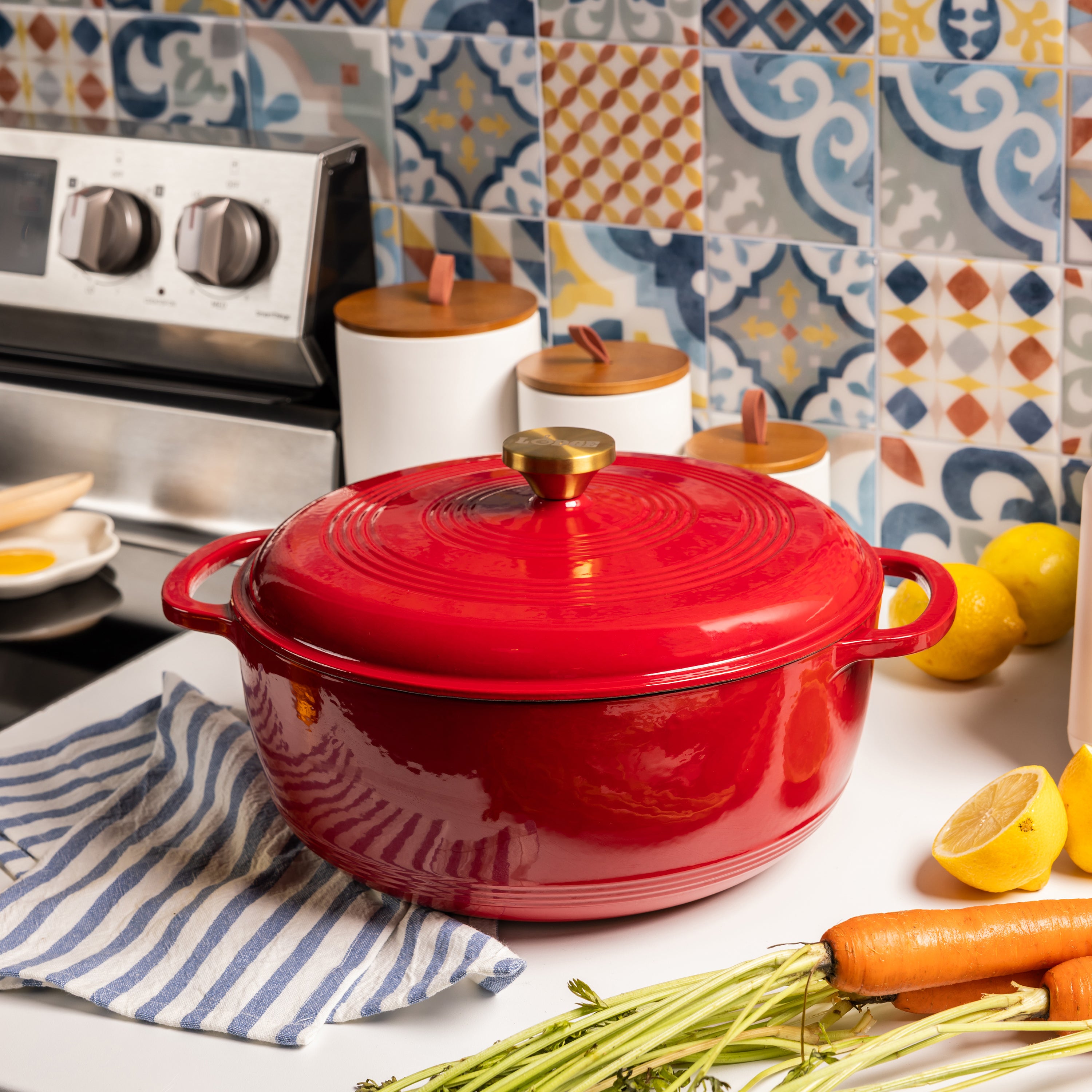 A glossy red enameled cast iron Dutch oven with a matching lid, set on a white countertop in a kitchen. The background features colorful patterned tiles, a stove, white canisters with wooden lids, lemons, carrots, and a blue-and-white striped cloth.