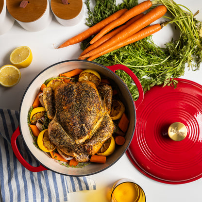 A red enameled cast iron Dutch oven with a lid, shown filled with a roasted whole chicken, carrots, and lemon slices. The Dutch oven is photographed from above on a white surface with carrots, lemon halves, and a striped cloth around it.
