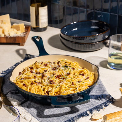 A dark teal cast iron skillet filled with creamy pasta and garnished with sun-dried tomatoes and parmesan, set on a blue fringed cloth on a kitchen counter with dining props.