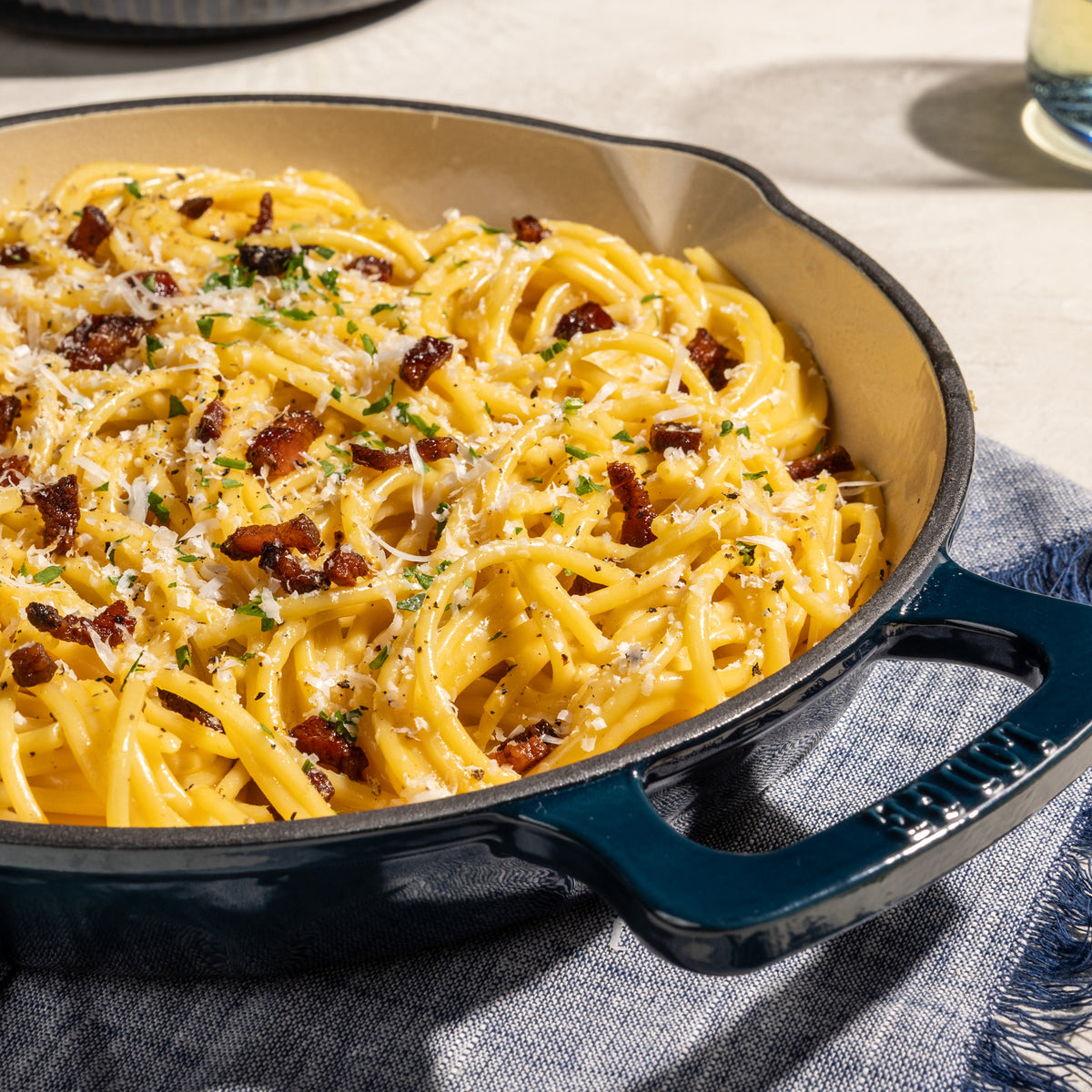 A close-up of creamy pasta carbonara in a dark teal enameled cast iron skillet, topped with crispy bacon bits, grated cheese, and chopped parsley, resting on a blue textured cloth.