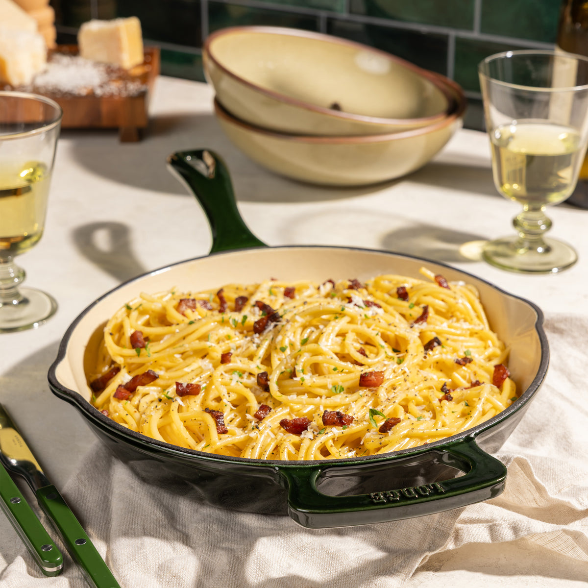 A black cast iron skillet with a dark green handle and interior, filled with creamy carbonara pasta topped with bacon bits and parmesan cheese, set on a dining table with wine glasses and bread in the background.
