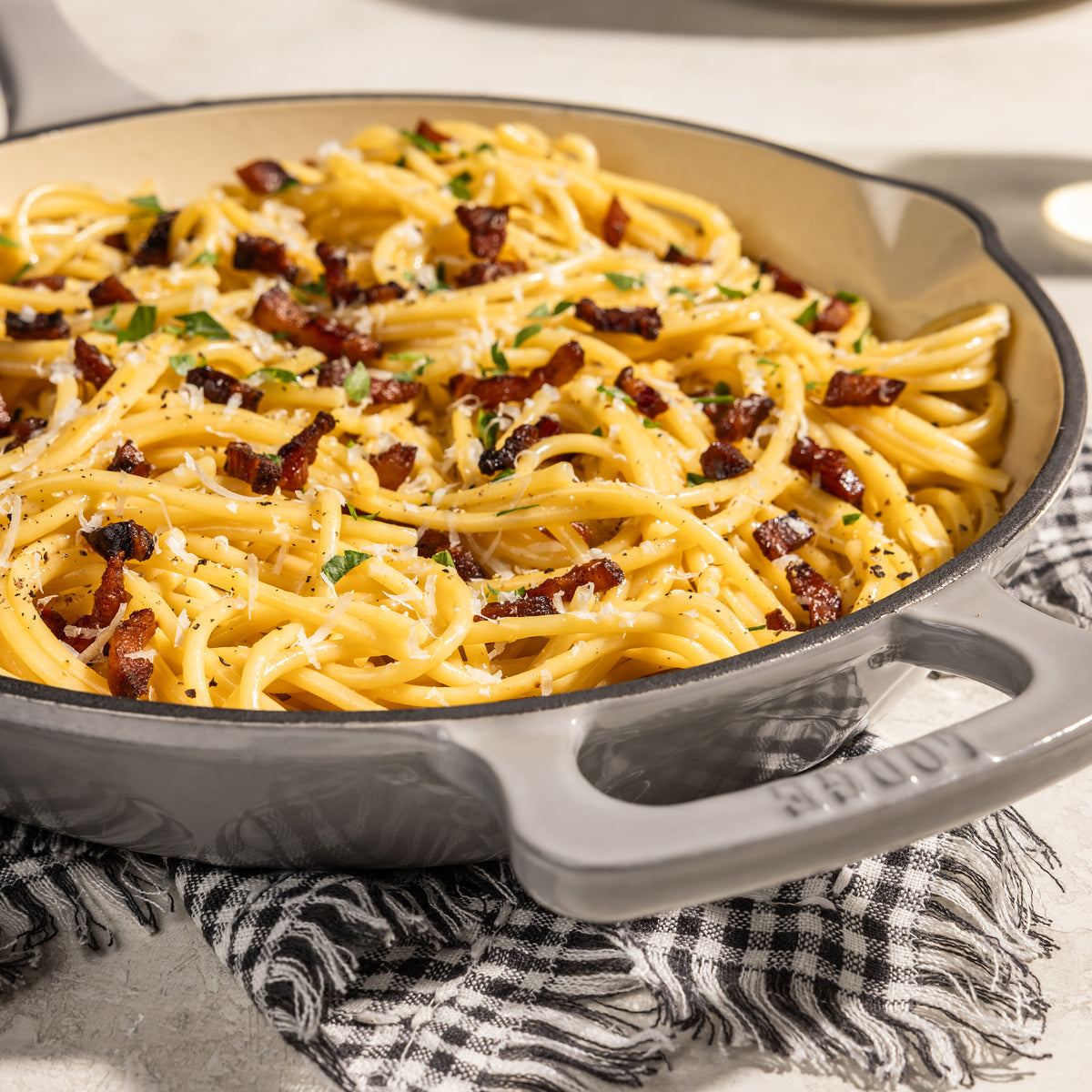 A close-up of a creamy pasta dish in a gray cast-iron skillet with bacon bits, herbs, and grated cheese, resting on a black-and-white checkered cloth.