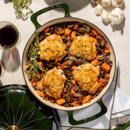 A top-down view of a green enameled cast iron Dutch oven filled with roasted chicken, carrots, and mushrooms, styled on a light stone surface with garlic, a glass of red wine, and herbs.