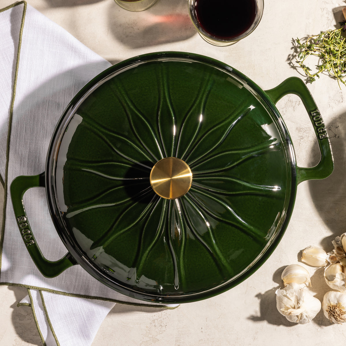 A dark green enameled cast iron pot with a matching lid, featuring a gold-tone central knob and embossed brand lettering on the handles, styled in a kitchen flat-lay with garlic, a glass of red wine, and herbs.
