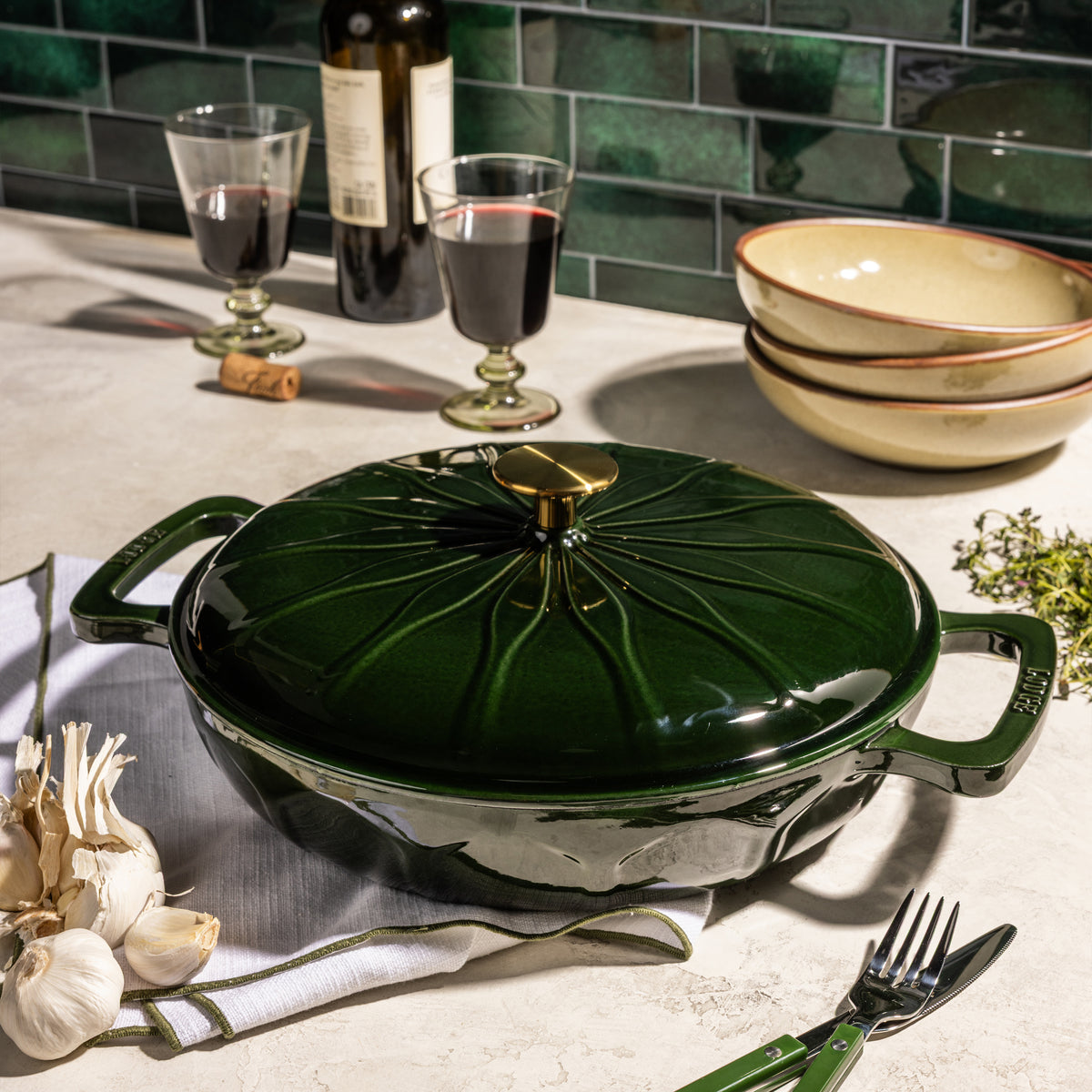 A dark green enameled cast iron casserole dish with a matching lid, styled on a kitchen countertop with wine glasses, garlic, a fork, and stacked bowls.