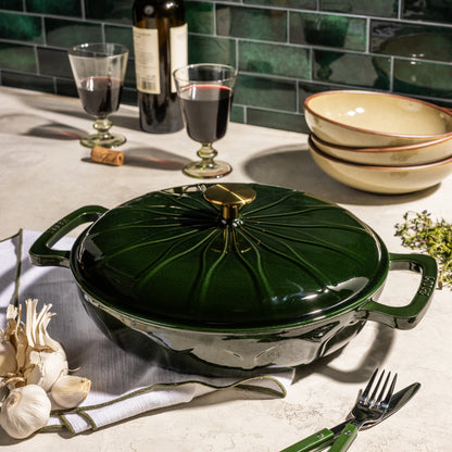 A dark green enameled cast iron casserole dish with a matching lid, styled on a kitchen countertop with wine glasses, garlic, a fork, and stacked bowls.