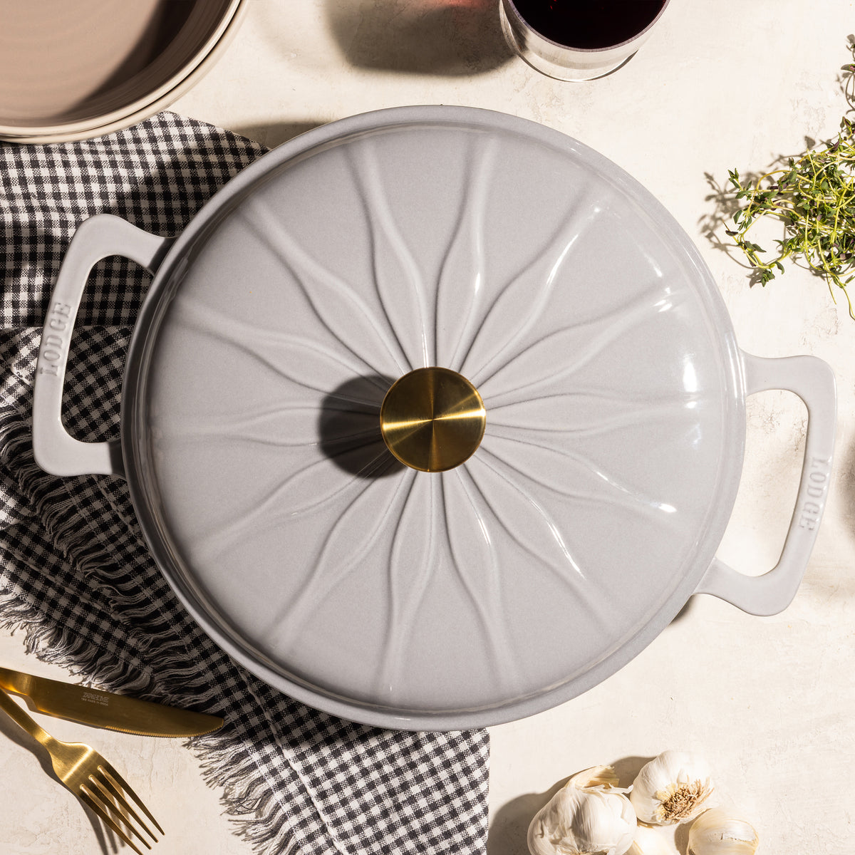 A light gray enameled cast iron pot with a matching lid featuring a sunburst pattern and a gold-tone knob, styled on a light beige surface with a black-and-white checkered cloth, garlic, herbs, and a gold fork.