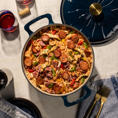 A top-down view of a dark blue enameled cast iron Dutch oven filled with a sausage and vegetable casserole, with its matching lid resting beside it on a light stone surface. The scene is styled with gold forks, a glass of red wine, and a bottle cork on a textured light gray background.