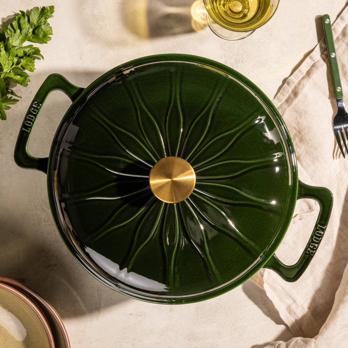 A dark green Lodge enameled cast iron pot with a lid, featuring a starburst pattern and a gold-tone knob, shown from a top-down angle on a beige linen surface with dining props.